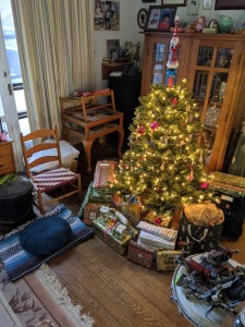 Picture of a small artisanal Christmas tree with lights and presents underneath, and to the left is a Zafu sitting on a folded blanket for a Zabuton.