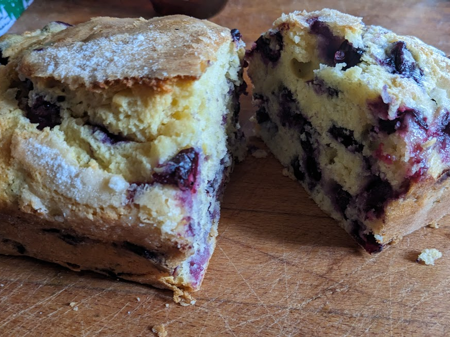A decorative photo of some yummy blueberry bread, which is sitting on a cutting board, and has had a couple of pieces sliced out from the center.