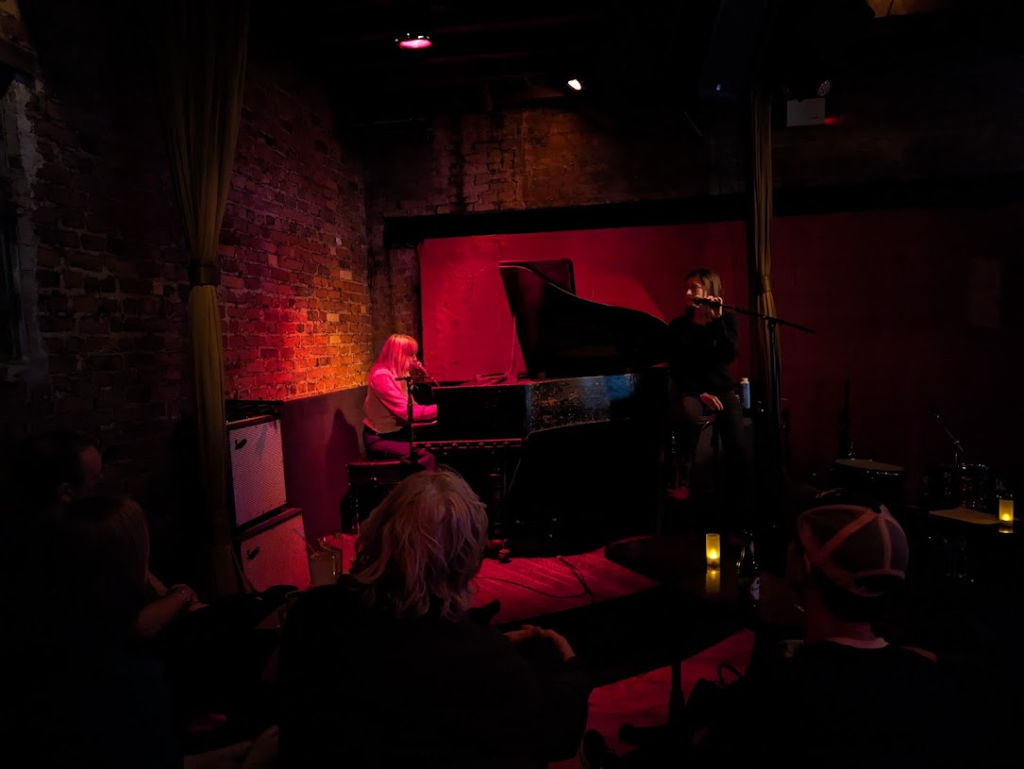 A dark club with exposed brick on the walls, a woman sitting at a piano and another woman on a stool beside the piano.