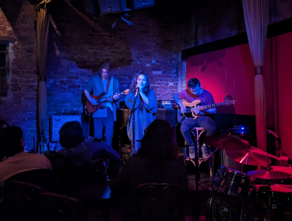 A dark club with exposed brick on the walls, a woman standing at a microphone with two guitarists behind her and a drummer off to the side.