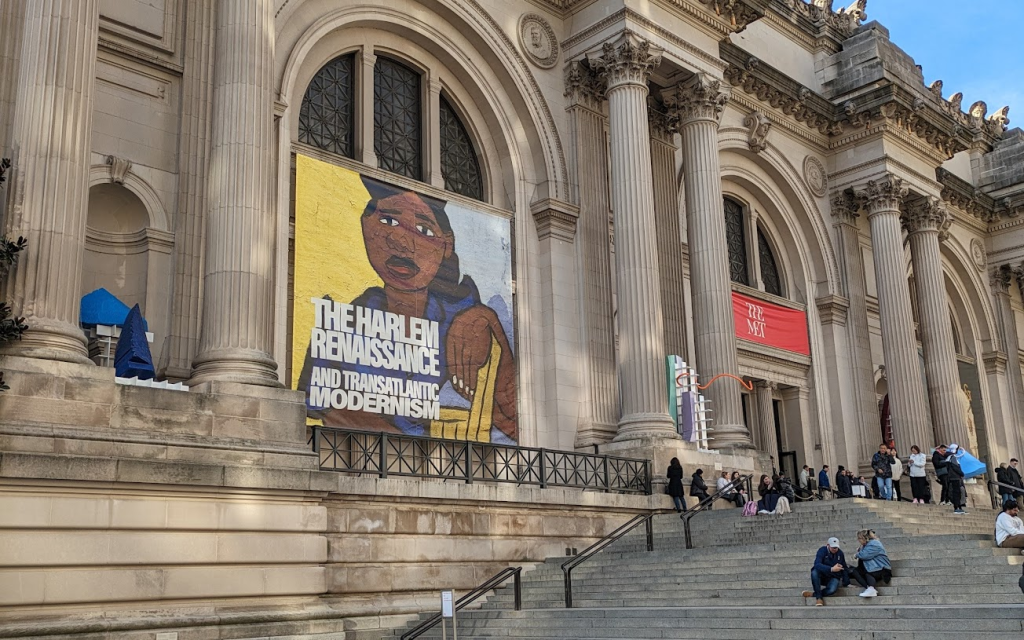 A photo of the front of the Metropolitan Museum of Art in New York City, with a large poster announcing The Harlem Renaissance and Transatlantic Modernism.